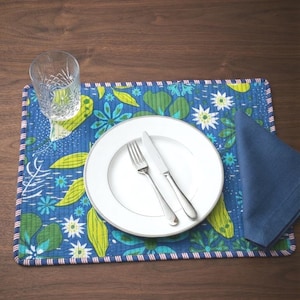 Puede incluir: Un mantel individual acolchado rectangular con un fondo azul vibrante con patrones florales verdes y azul claro, bordeado por un ribete a rayas rojas y blancas. Un plato llano blanco, un tenedor de plata, un cuchillo de plata, una servilleta azul y un vaso.
