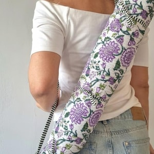 May include: A cylindrical yoga mat bag featuring a white base with a repeating purple floral pattern and green stems. The bag includes a black and white striped fabric shoulder strap and matching striped trim along the top and bottom edges.
