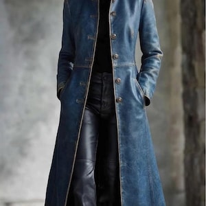 May include: A long, distressed blue leather coat featuring a high collar, a front button closure with metallic buttons, and two side pockets. The coat is worn over a black turtleneck sweater and black leather trousers, creating a layered look.
