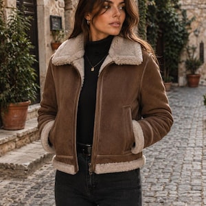 May include: A brown suede jacket with a cream-colored faux shearling lining, a wide fold-over collar, long sleeves with shearling cuffs, a front zipper closure, and two side pockets, worn over a black turtleneck sweater and black jeans.