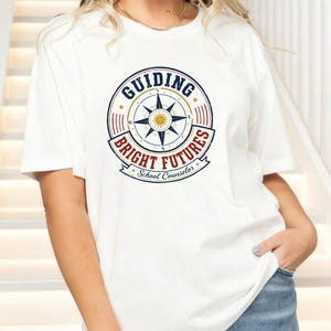 May include: A white short-sleeved t-shirt featuring a circular graphic with a compass rose in the center. The text reads "GUIDING BRIGHT FUTURES" in blue and red, with "School Counselor" in a banner below the compass design.