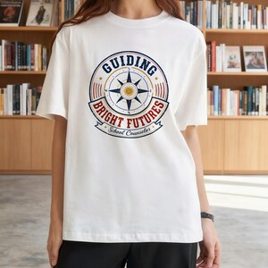 May include: A white short-sleeved t-shirt featuring a circular graphic on the chest. The design includes a central compass rose, the text "GUIDING BRIGHT FUTURES" in blue and red, and the words "School Counselor" at the bottom in blue.