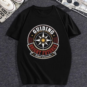 May include: A black short-sleeve t-shirt featuring a circular graphic on the chest. The design includes a compass rose, a central sun icon, and the text "GUIDING BRIGHT FUTURES" with "School Counselor" written in a banner at the bottom.