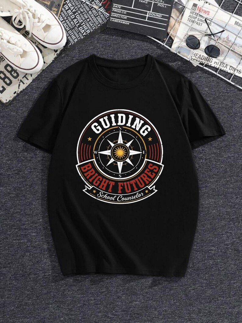 May include: A black short-sleeve t-shirt featuring a circular graphic on the chest. The design includes a compass rose, a central sun icon, and the text "GUIDING BRIGHT FUTURES" with "School Counselor" written in a banner at the bottom.