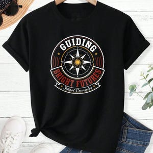 May include: A black short-sleeve t-shirt featuring a circular graphic with a compass rose in the center. The text reads "GUIDING BRIGHT FUTURES" in red and white, with "School Counselor" printed in white script at the bottom of the design.