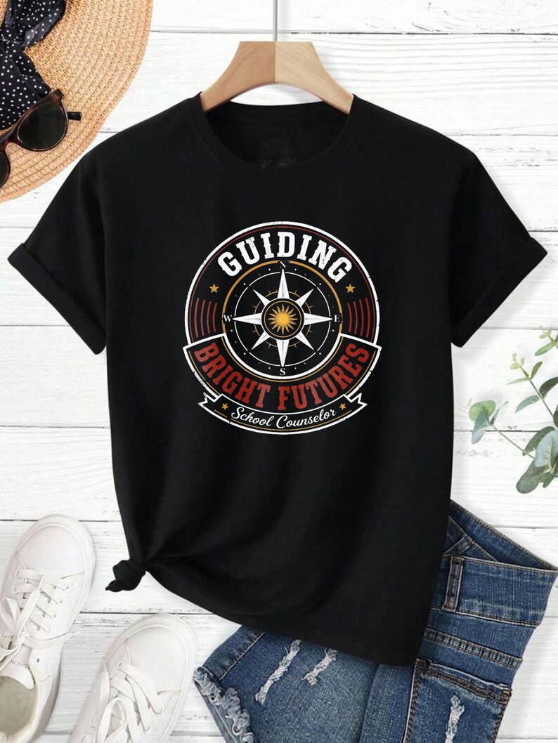 May include: A black short-sleeve t-shirt featuring a circular graphic with a compass rose in the center. The text reads "GUIDING BRIGHT FUTURES" in red and white, with "School Counselor" printed in white script at the bottom of the design.