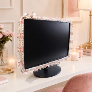 May include: A computer monitor frame cover featuring a white lace border adorned with small pink fabric roses and a matching satin bow at the top centre. The decorative trim surrounds the black screen, providing a soft, textured aesthetic.