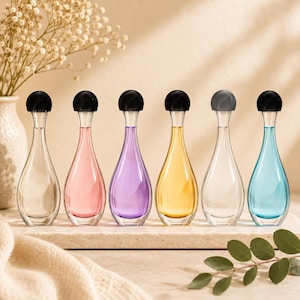 May include: Six glass perfume bottles with teardrop-shaped bodies and narrow necks, each topped with a round black cap. The bottles contain liquids in clear, pink, purple, yellow, clear, and blue colors, arranged in a row on a flat stone surface.