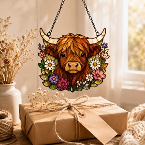 May include: A stained glass suncatcher featuring a brown Highland cow head surrounded by colorful flowers including white daisies and purple blossoms. The piece is suspended by a black metal chain and features detailed leaded glass lines.