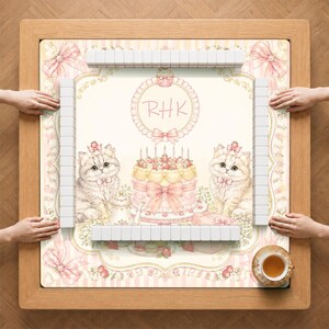 May include: A square mahjong table with a light pink and cream striped surface featuring two fluffy kittens, a tiered strawberry cake, a teapot, and the letters RHK in a circular frame. White mahjong tiles are arranged in four rows on the table.