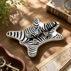 Può includere: Un piccolo vassoio in ceramica dalla forma irregolare a stella, caratterizzato da un motivo a strisce zebrate bianche e nere con un bordo dorato. Quattro dadi poliedrici metallici a più facce poggiano all'interno del vassoio.