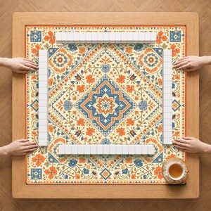 May include: A square wooden game table featuring a patterned mat with orange and blue floral motifs, geometric borders, and small beetle illustrations. Four rows of white rectangular mahjong tiles are arranged in a square formation.