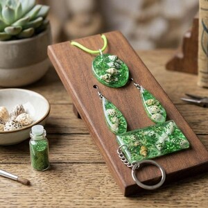 Può includere: Un set di gioielli composto da una collana, due orecchini a goccia e un portachiavi rettangolare, realizzati in resina verde con piccole conchiglie bianche incorporate. La collana presenta un cordino verde neon e il portachiavi un anello in metallo argentato.