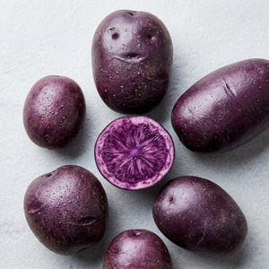 Puede incluir: Un conjunto de seis patatas moradas dispuestas en un patrón circular. Cinco patatas están enteras con piel lisa de color morado oscuro, y una patata está cortada por la mitad en el centro, revelando un interior morado intenso con un patrón estelar.