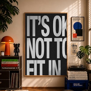 May include: A large rectangular wall art print in a wooden frame featuring the white text IT'S OK NOT TO FIT IN against a textured black background. The poster is displayed on a wall alongside other framed art prints and various home decor items.