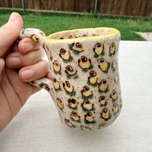 May include: A ceramic mug with a speckled cream-colored glaze featuring a repeating pattern of small yellow and brown rose motifs. The mug has a thick, uneven rim painted in bright yellow and a sturdy handle with matching floral details.