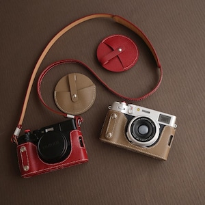May include: Two digital cameras with textured leather cases, one in red and one in tan, featuring silver metal snap closures. A long red leather camera strap is attached to the red camera, accompanied by two circular leather lens caps in red and tan.