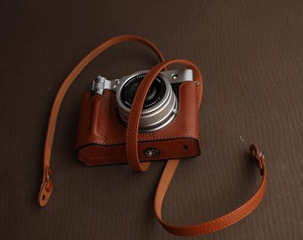 以下が含まれることがあります： タンカラーのレザー製保護ケースに入ったシルバーのデジタルカメラ。同色のレザーストラップ付き。カメラには円形のレンズ、ダイヤル操作が可能なシルバーのトッププレート、質感のあるレザー仕上げのボディが備わっています。