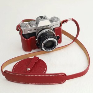 Puede incluir: Una cámara Fujifilm X-T20 plateada con un objetivo Voigtlander negro, dentro de una funda de cuero rojo texturizado con costuras blancas. Una correa de cuero rojo a juego y una tapa de objetivo circular roja están unidas al cuerpo.