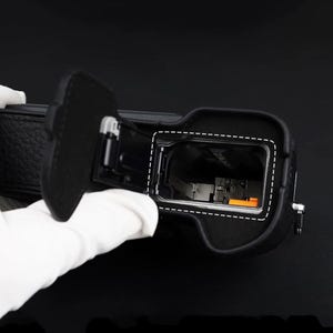 以下が含まれることがあります： 開いたヒンジ付きドアを備えた黒いカメラのバッテリーコンパートメント。内部の空洞、金属製のスプリング接点、小さなオレンジ色のリリースレバーが見えます。コンパートメントエリアは、カメラ本体の白い破線で囲まれています。