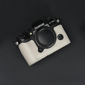 以下が含まれることがあります： 黒いFujifilm X-T50デジタルカメラボディと、質感のあるライトグレーのレザー調ハーフケース。カメラにはFX mountと記された中央のレンズマウントキャップ、上部のダイヤル、保護ケース前面の小さな円形ボタンが備わっています。
