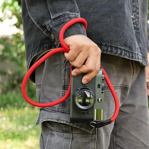 May include: A dark gray digital camera with a prominent circular lens and a bright red rope wrist strap. The camera body features a textured finish and a small rectangular window on the front. The red strap is looped around a wrist for secure holding.
