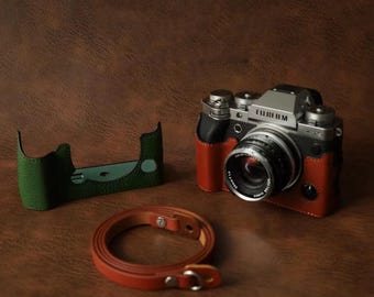 以下が含まれることがあります： シルバーとブラックの富士フイルム製カメラ。タンカラーのレザーハーフケース、別のグリーンのレザーハーフケース、お揃いのタンカラーのレザーストラップが付属しています。レンズには35mm F1.4という文字が刻まれています。