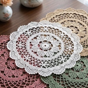 May include: A set of four round crochet doilies in white, dusty pink, sage green, and beige. Each doily features an intricate, circular floral pattern with a scalloped edge, showcasing detailed needlework and a delicate, openwork lace design.