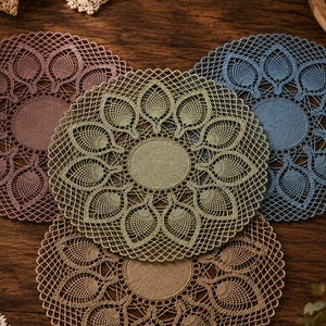 May include: Four circular lace doilies in mauve, sage green, dusty blue, and tan. Each doily features a solid circular center surrounded by a detailed, repeating leaf-like pattern and an intricate outer lattice border made of fine thread.