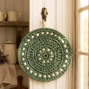 May include: A circular, hand-crocheted trivet featuring a deep sage green yarn base with a contrasting cream-colored woven accent detail. The round mat is suspended from a small, antique-style brass hook attached to a vertical white wooden panel.