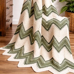 May include: A crocheted blanket featuring a chevron pattern in alternating bands of cream and sage green. The textile displays a textured stitch design with a zigzag edge, showcasing a repeating geometric motif throughout the entire piece.