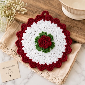 May include: A round, crocheted potholder featuring a scalloped deep red border, a wide white circular center, and a central floral motif with green leaves and a deep red flower. A small loop at the top allows for hanging this kitchen textile.