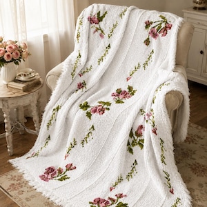 May include: A white knitted throw blanket featuring a repeating floral pattern of pink roses and green leaves. The blanket has a textured, ribbed knit construction with a fringed edge, draped over a piece of furniture.