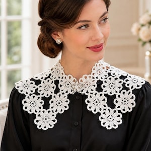 May include: A white crocheted collar featuring a repeating floral pattern made of interconnected circular motifs. The collar is secured at the center front with a small, silver-toned star-shaped brooch and is worn over a solid black long-sleeved shirt.