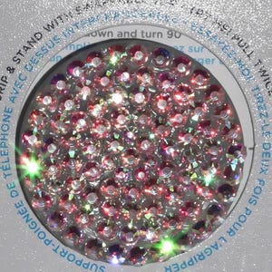 May include: A circular phone grip and stand featuring a dense arrangement of small, multifaceted pink and iridescent crystals. The surrounding packaging includes text that reads "GRIP & STAND WITH SWAPPABLE TOP" and "TRY ME! PULL TWICE".
