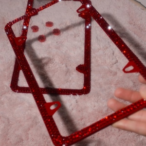 May include: Two rectangular license plate frames covered in small, reflective red rhinestones. Each frame features four mounting holes with red metal tabs. The surface of the frames is densely packed with sparkling red crystals that catch light.