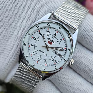 May include: A silver-tone Seiko 5 automatic wristwatch with a white dial, featuring a mesh metal strap, a day-date window at the three o'clock position, and a dual-scale display with 12-hour and 24-hour markers, marked with Japan Made text.