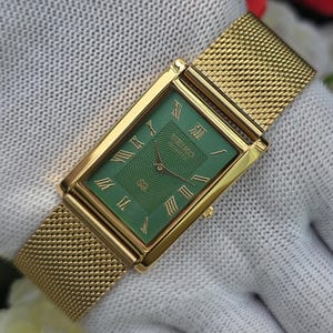 May include: A rectangular gold-tone wristwatch featuring a green dial with a wavy pattern, gold Roman numeral hour markers, and gold hands. The watch has a gold-tone mesh metal strap and the Seiko Quartz logo is printed on the dial face.
