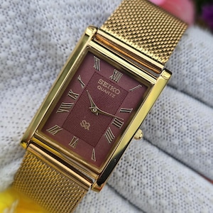 May include: A rectangular gold-tone wristwatch with a mesh band and a deep red dial. The dial features a textured wave pattern, gold-tone hands, and gold Roman numeral hour markers. The text Seiko Quartz is printed in gold on the upper dial.