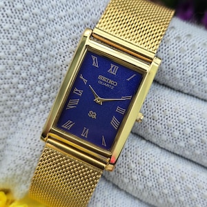 May include: A rectangular gold-tone Seiko Quartz watch with a deep blue textured dial. The watch features gold-colored Roman numeral hour markers, gold hands, and a gold-tone mesh metal strap. The brand name Seiko and Quartz are printed in white.