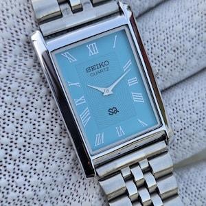 Może przedstawiać: Prostokątny zegarek Seiko Quartz z jasnoniebieską tarczą o subtelnym falistym wzorze, srebrnymi rzymskimi cyframi i srebrnymi wskazówkami. Zegarek posiada polerowaną bransoletę z ogniw w kolorze srebrnym z bezpiecznym zapięciem.