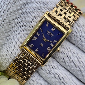 Puede incluir: Un reloj de pulsera rectangular Seiko Quartz en tono dorado con esfera azul profundo, números romanos dorados, manecillas doradas y una correa de eslabones texturizados. La esfera muestra el logotipo de Seiko Quartz y un pequeño emblema.