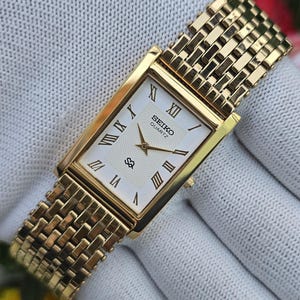May include: A gold-tone rectangular Seiko Quartz wristwatch featuring a white dial with black Roman numeral hour markers, gold hands, and a textured center pattern. The watch includes a matching gold-tone metal link bracelet with a woven design.
