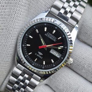May include: A silver-tone Citizen Automatic wristwatch with a black dial, featuring a fluted bezel, a stainless steel link bracelet, luminous hands, a red second hand, and a day-date window displaying WED 24. The dial includes the text Citizen Automatic and 21 Jewels.
