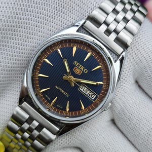 May include: A silver-tone stainless steel Seiko 5 automatic wristwatch with a dark blue dial featuring a textured striped pattern, gold-tone hands and hour markers, and a day-date window at the three o'clock position. The watch has a metal link band.