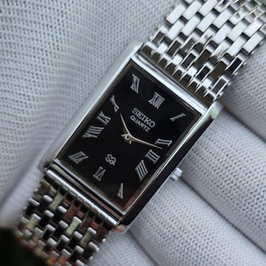 May include: A rectangular Seiko Quartz wristwatch with a black dial, silver-tone Roman numeral hour markers, and silver hands. The watch features a polished silver-tone metal case and a matching multi-link bracelet with a textured, woven appearance.