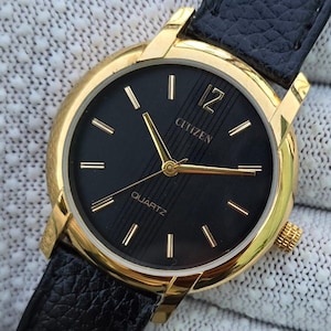 May include: A Citizen quartz wristwatch featuring a round gold-tone case, a black dial with vertical textured stripes, gold-tone hands and hour markers, a prominent numeral 12, and a black textured leather strap with a gold-tone crown.