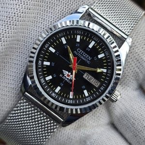 May include: A Citizen automatic wristwatch with a black dial, silver-tone hands, and a red second hand. The watch features a fluted bezel, a stainless steel mesh strap, and a day-date window at the three o'clock position. The dial displays 21 jewels.