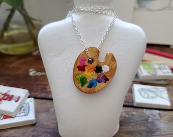 Può includere: Collana con pendente a forma di tavolozza da artista in legno con catena argentata. La tavolozza è dipinta con tocchi di vari colori, tra cui rosa, giallo, verde, blu e viola. La collana è esposta su un busto di manichino bianco.