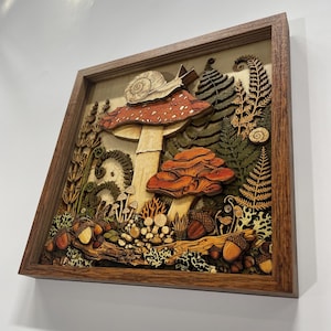 May include: A multi-layered wooden wall art piece in a dark brown frame featuring a detailed forest scene. The composition includes a large red-capped mushroom with a snail on top, smaller orange mushrooms, ferns, acorns, and moss, all rendered in wood.